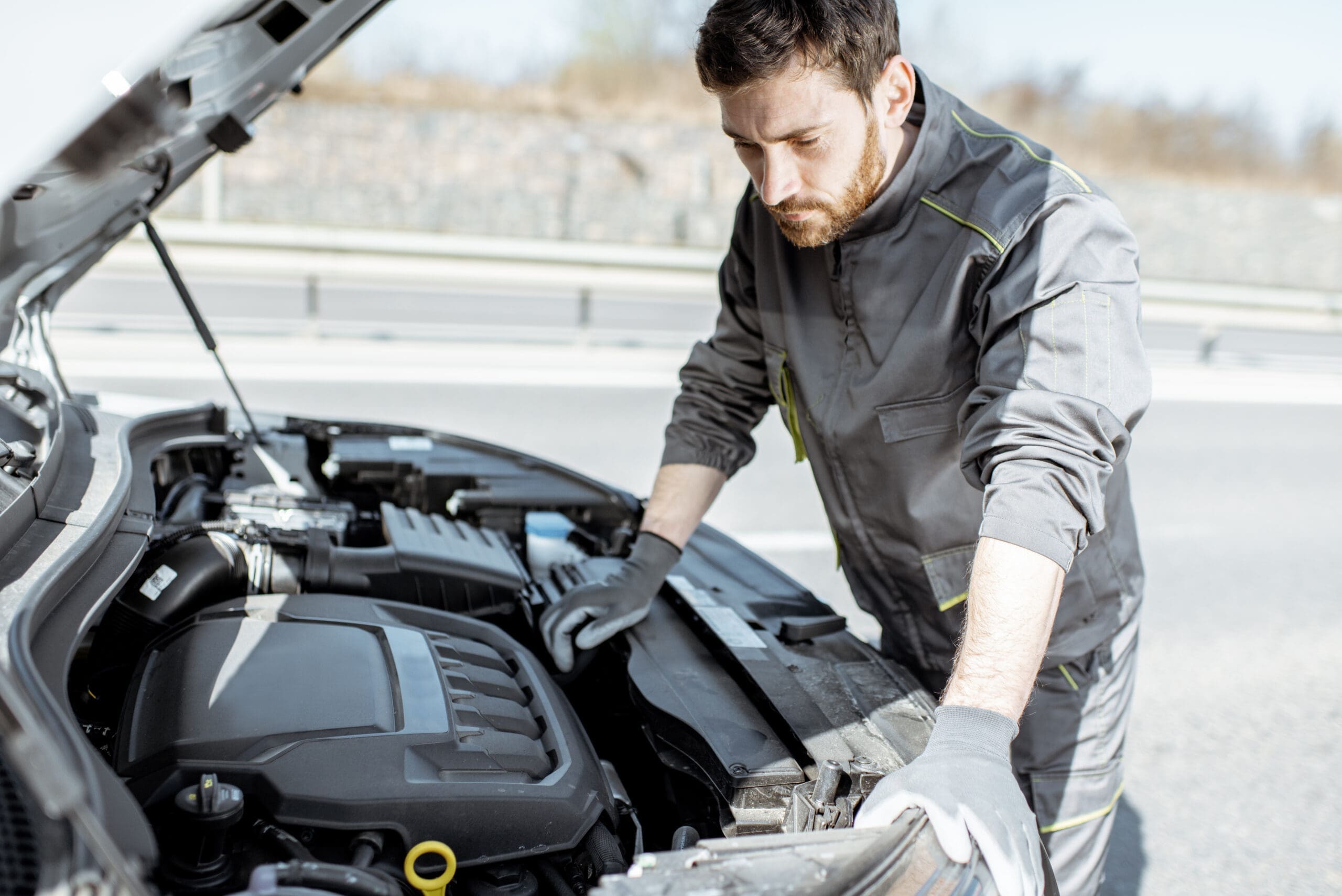 Handsome auto mechanic or road assistance worker in uniform repairing engine of the broken car on the road