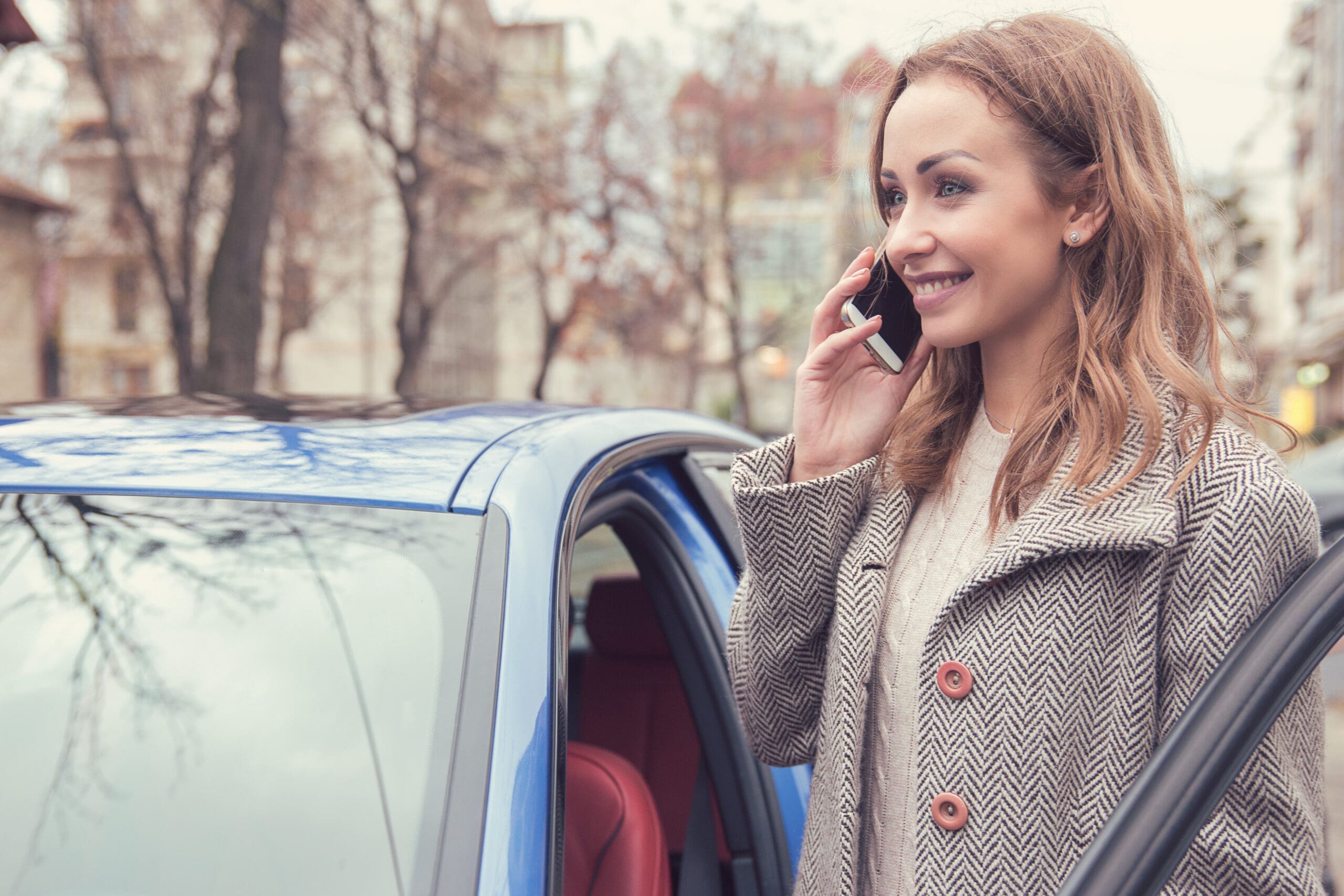 Pretty woman in outerwear having phone call standing on street near automobile and smiling.